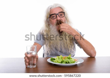 Studio shot of senior bearded man wearing eyeglasses while eating plate of salad with glass of water on wooden table