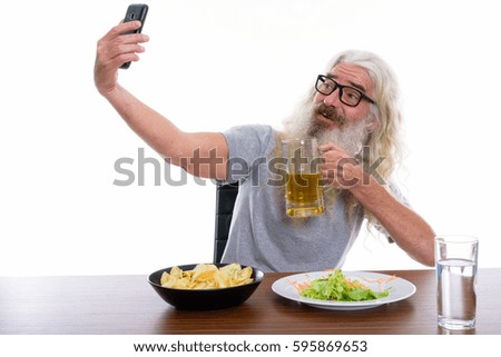 Happy senior bearded man smiling while taking selfie picture with mobile phone while holding glass of beer with healthy and unhealthy foods on wooden table