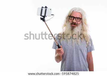 Studio shot of happy senior bearded man smiling while wearing eyeglasses and taking selfie picture with mobile phone on selfie stick