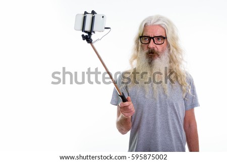 Studio shot of senior bearded man wearing eyeglasses and taking selfie picture with mobile phone on selfie stick