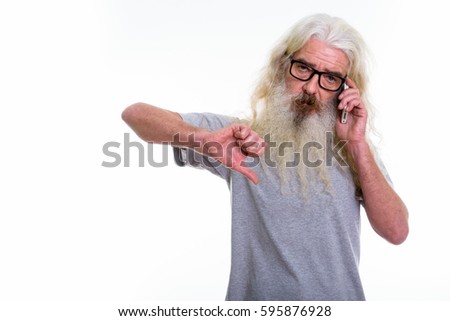 Studio shot of senior bearded man giving thumb down while talking on mobile phone