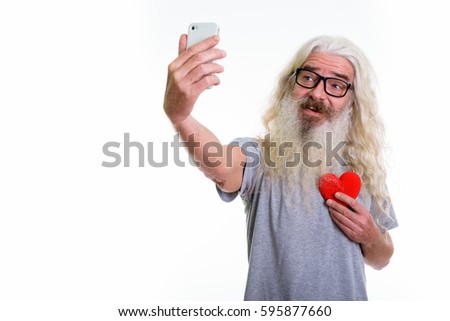 Studio shot of happy senior bearded man smiling and holding red heart while taking selfie picture with mobile phone