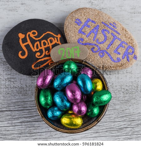 Happy Easter 2017 lettering written on pebbles with chocolate eggs wrapped in colorful foil. Square image