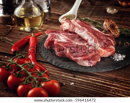 Raw pork neck steak on a stone plate. Salt shaker, pepper shaker. Roasted Garlic. Hot chili pepper. Rosemary.