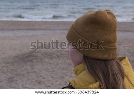 A European girl with long blond long hair in a brown hat and a mustard-colored jacket standing on the beach and looking at the sea
