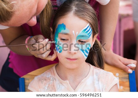 Child animator, artist's hand draws face art to little girl. Blue butterfly painting. Children birthday party entertainment