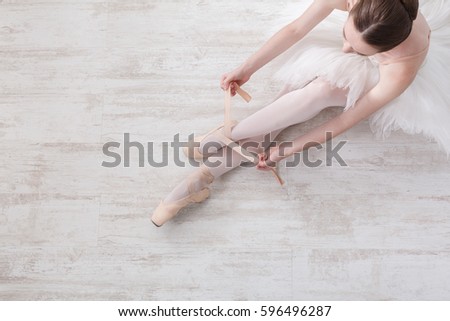 Beautiful legs of young ballerina who puts on pointe shoes at white wooden floor background, top view from above with copy space. Ballet practice. Beautiful slim graceful feet of ballet dancer.
