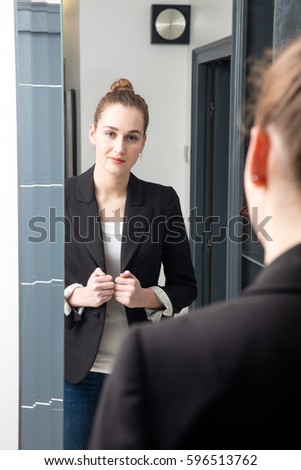 successful beautiful young woman with winning body language standing in front of her mirror, getting ready for business from home, looking at her reflection in the background