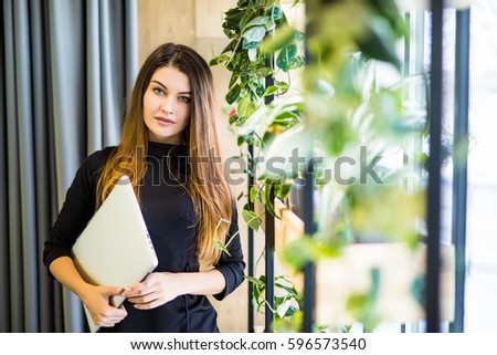 Portrait Of Successful Smiling Businesswoman Holding Laptop Computer In Her Hands. Happy Beautiful Confident Woman Standing At Office Wall With Notebook. Business People.