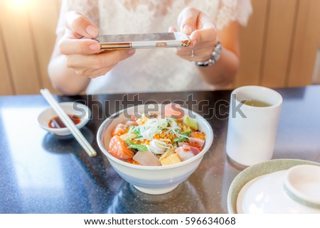 Woman's hands taking picture of Japanese sashimi dish by smartphone camera, soft-focus on hand and defocus in background.