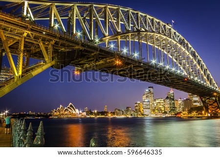 Sydney harbour bridge at sunset with bright illumination of steel arch and columns reflecting in the blurred waters of harbour with Sydney city CBD in the background