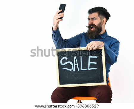Bearded man, long beard. Brutal caucasian amused hipster with moustache making selfie on cell phone and holding sale inscription on blackboard, sitting on the chair isolated on white background