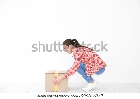 Posture concept. Young woman lifting heavy cardboard box against white wall background Royalty-Free Stock Photo #596816267
