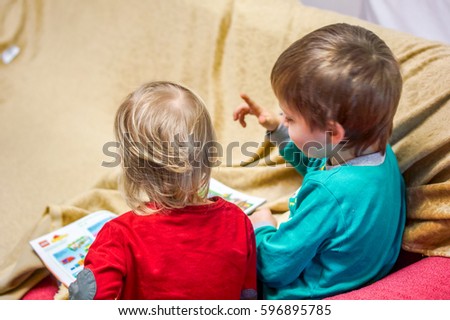 two small kids brother and sister sit on soft burgundy red sofa and reading book with pictures back view. Older brother show to small blonde sister pictures on soft book in a homely atmosphere