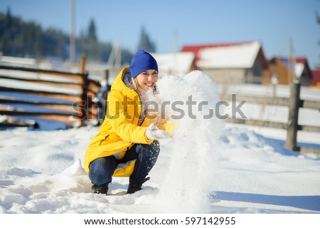 Young beautiful woman cheerfully throws snow. Fine winter day. Ground is covered by snow. Woman in a bright yellow down-padded coat rejoices to snow. On her face a happy smile. Beauty rural areas.