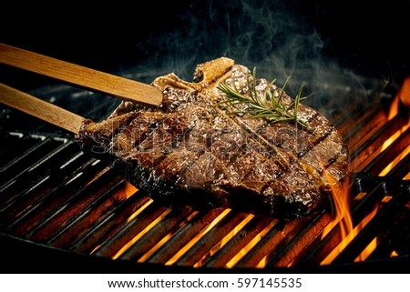 Grilling a tasty tender marinated t-bone steak seasoned with fresh rosemary on a barbecue fire with hot fiery coals in a close up view Royalty-Free Stock Photo #597145535