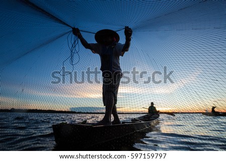 Thai fisherman on wooden boat casting a net for catching freshwater fish in nature river in the early evening before sunset
