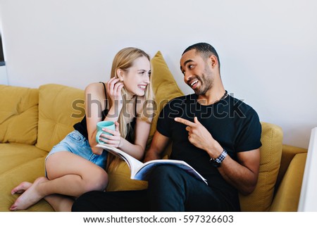 Couple joking, talking, having fun, smiling on the couch at home. Lovers relax, read a magazine, drinking tea. Positive mood, conversation, teasing.