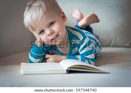 Little, pensive, lost in thought, cheerful and dreamy two years old boy lying on his belly on the sofa, with open book in front of him.