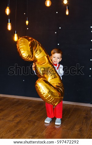Little boy in white shirt, red pants and bow-tie having fun on his birthday and playing with big, gold balloon digit. Fun and happy child on the background of black wall and gold Edison lamps