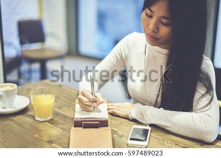 Pensive young female artist resting during weekends in coffee shop listening to favorite music via smartphone and earphones while making creative sketch drawings in notepad inspiring by songs