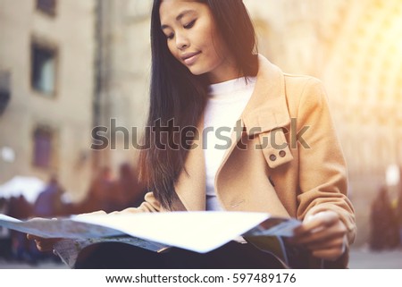 Charming concentrated young hipster girl spending weekends traveling alone in old European town using map to navigate in city streets sitting outdoors resting and planning next touristic attraction