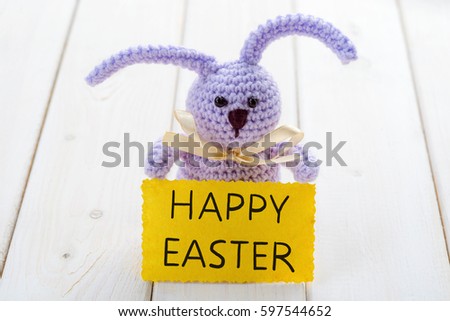 Lilac knitted bunny holding a yellow Happy Easter card, close-up. Light wooden background. Spring Christian traditions.
