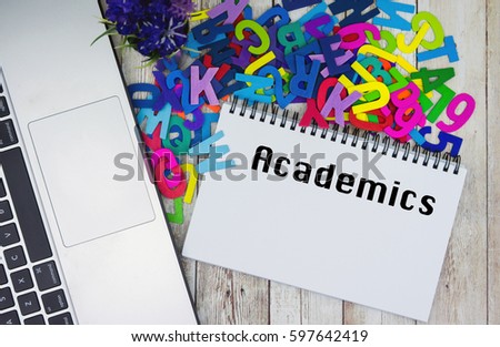 ACADEMICS text on notebook with laptop and piles of alphabet on wooden table. 