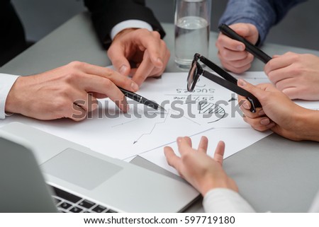Discussing new development strategy. Negotiation participants make depictions and have a lively discourse. There are papers with drafts, a notebook and a glass of water on the table. Royalty-Free Stock Photo #597719180
