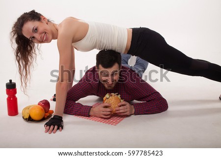 Working her core muscles. Full length of young beautiful woman in sportswear doing plank while a man eats a burger. Problem of choice. Healthy and unhealthy food. 