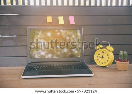 A wooden desk with laptop,yellow clock,cactus in a pot and multicolor note pad  in morning,working space and working out.