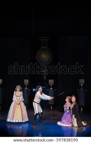 Four actors play roles on stage, a young man in an old frock coat, an elderly man in military uniform and wearing a cocked hat and two female actresses in medieval dresses with wide skirts