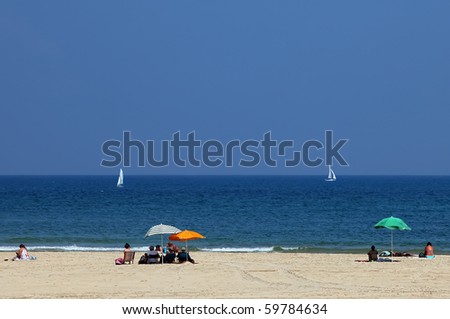 People sitting under umbrellas on a sandy sea beach and looking at white yachts
