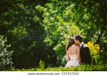 Look over green bushes at groom kissing bride's head tender while they stand among green bushes