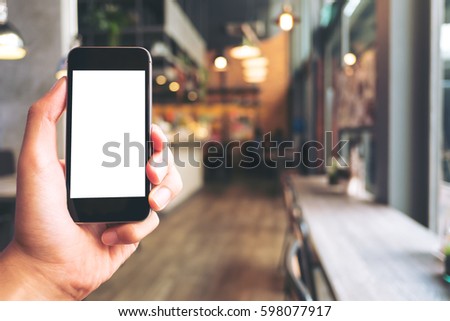 Mockup image of hand holding black mobile phone with blank white screen in modern cafe