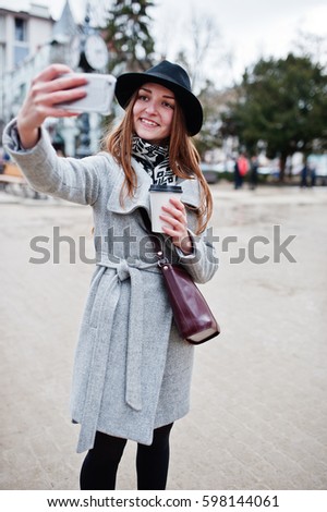 Young model girl in a gray coat and black hat with leather handbag on shoulders stay with plastic cup of coffee and making selfie at street of city.