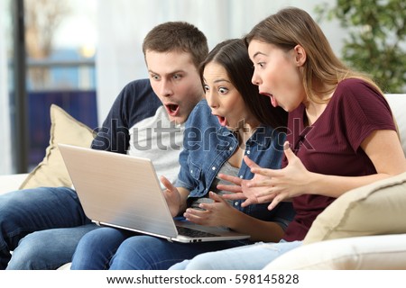 Three amazed friends watching media content on line in a computer sitting on a couch in the living room at home Royalty-Free Stock Photo #598145828