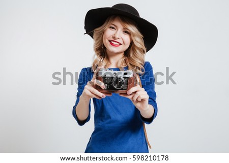 Picture of pretty young woman dressed in blue dress wearing hat holding camera and posing over white background.