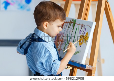 Cute, serious and focused, three years old boy in blue shirt and jeans apron drawing on canvas standing on the easel. Concept of early childhood education, painting, talent, happy family or parenting