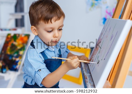 Cute, serious and focused, three years old boy in blue shirt and jeans apron drawing on canvas standing on the easel. Concept of early childhood education, painting, talent, happy family or parenting Royalty-Free Stock Photo #598212932
