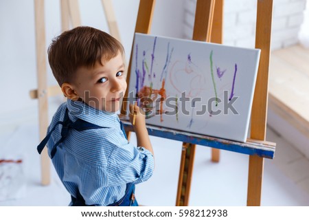 Close-up portrait of cute, smiling, white three years old boy in blue shirt and jeans apron with brush in the hand. Concept of early childhood education, painting, talent, happy family or parenting Royalty-Free Stock Photo #598212938