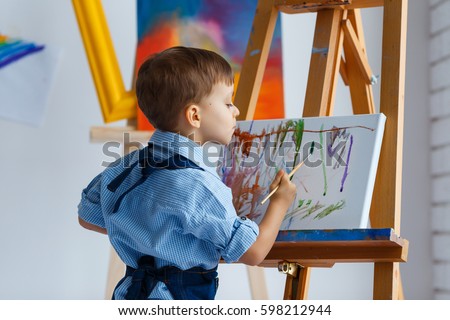 Cute, serious and focused, three years old boy in blue shirt and jeans apron drawing on canvas standing on the easel. Concept of early childhood education, painting, talent, happy family or parenting Royalty-Free Stock Photo #598212944