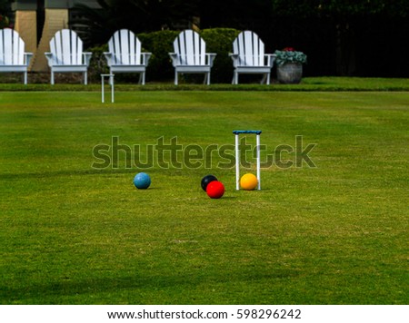 Adirondack chairs sit behind a croquet court where multiple balls are being played around a hoop or wicket on a relaxing summer day. Royalty-Free Stock Photo #598296242