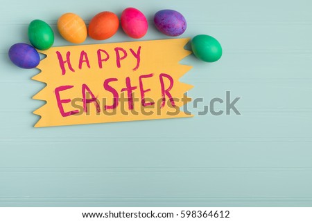 Dyed Easter Eggs Above a Yellow Sign in Bright, Cheerful Colors.  Happy Easter is Hand Painted on the sign with Wood Board Background in Light Teal Blue and Room or Space for Copy, Text or Your words