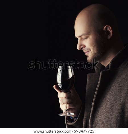 Handsome young bald man smelling red wine before tasting and drinking it. Man wearing spring coat and holding a glass of red wine in his hand. Low key portrait
