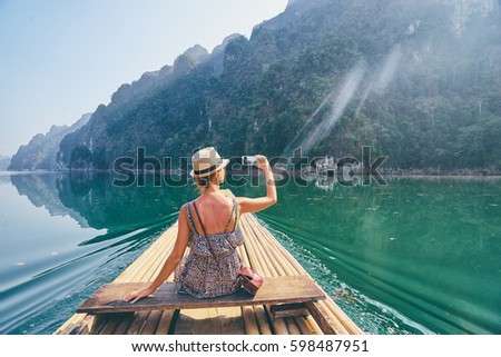 Travel and technology. Pretty young woman taking photo on smartphone sailing National Park lake on traditional boat.
