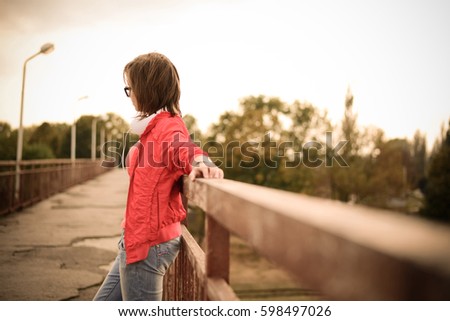 A portrait of a girl standing on a bridge, an old emergency bridge, fashionable clothes, stylish glasses and headphones, a hairstyle, a leather bracelet on her arm, a sunset