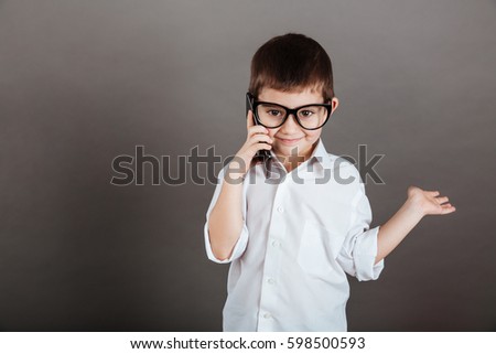 Happy little boy in glasses talking on mobile phone and holding copyspace on palm over grey background