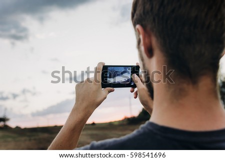 man hipster traveler taking photo holding smart phone, of beautiful sunset scenery in summer field. instagram photography. exploring and discovering. space for text. lifestyle