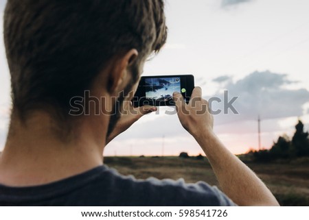 man holding phone and taking photo of amazing sunset landscape view in summer field. travel and vacation. exploring and discovering nature. instagram photography. space for text